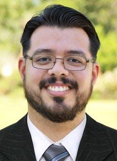 Man with dark hair, wearing glasses, and wearing black blazer and gray tie, standing in front of greenery.