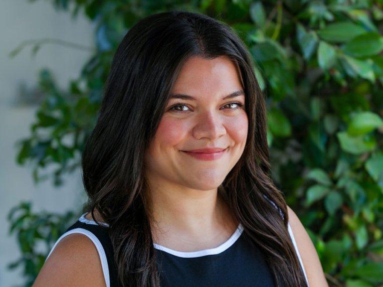 Close-up headshot of Emily Butts, who has long wavy dark hair and stands in front of a green ivy wall.